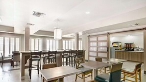 A modern dining area with multiple tables and chairs, a kitchen area with appliances, and large windows with blinds.