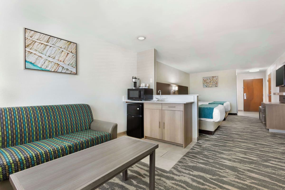 A compact hotel room with a kitchenette, a sofa, a bed, and a small table.