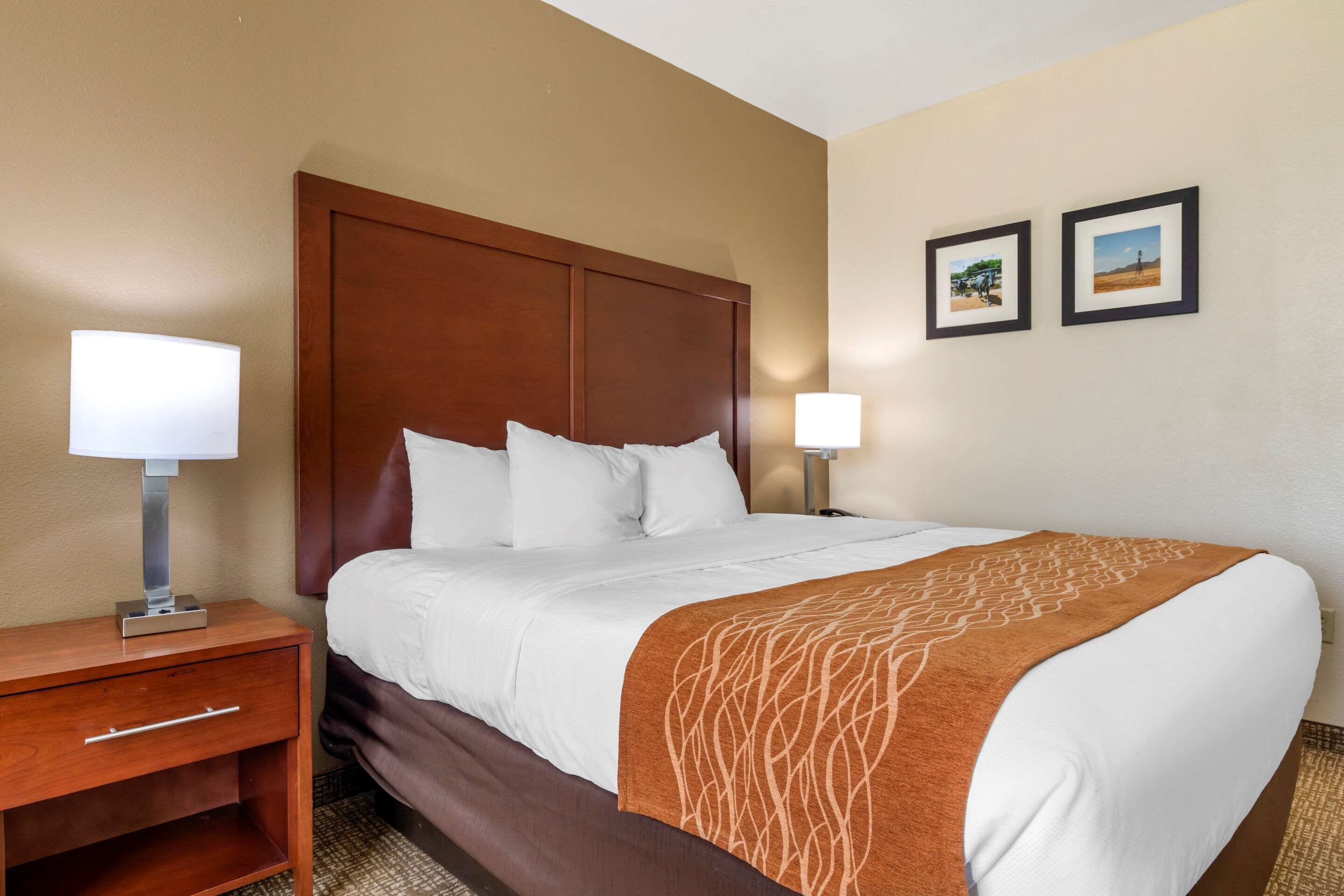 A hotel room with a large bed, wooden headboard, two bedside lamps, a nightstand, and two framed pictures on the wall.