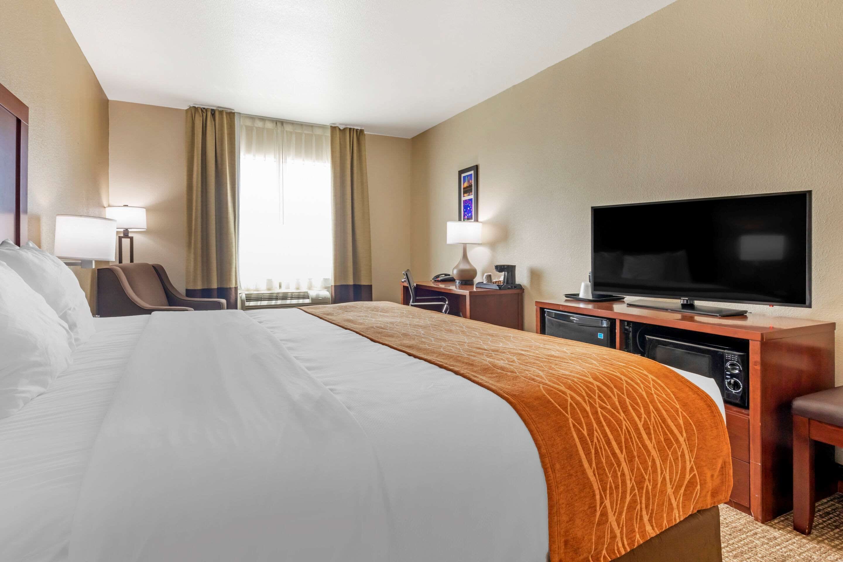 A hotel room with a large bed, a desk with a television, a chair, and a lamp.