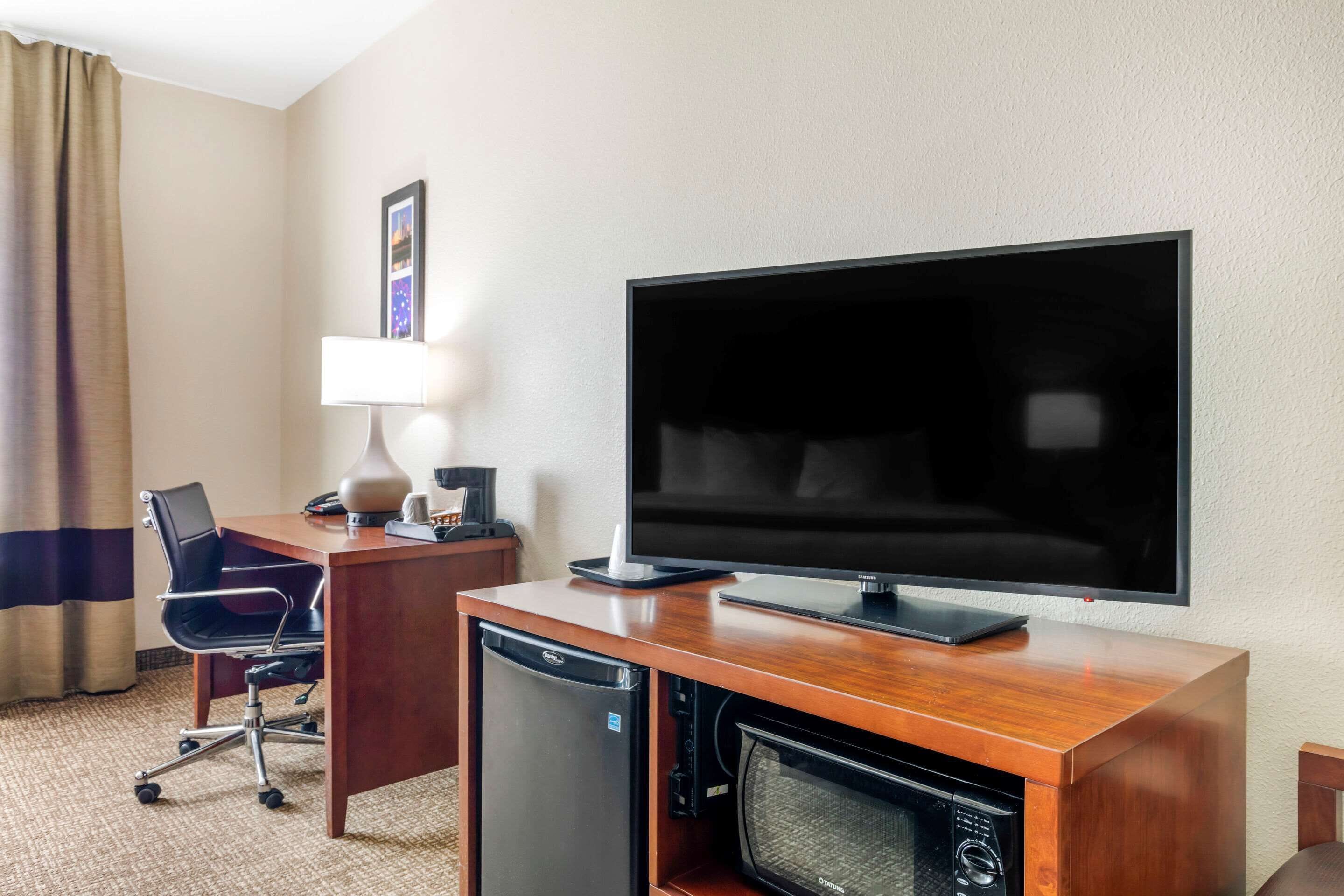 Hotelkamer met een houten bureau, een flatscreen televisie, een compact magnetron, een minibar, een zwarte bureaustoel, een lamp en een lijst met een afbeelding aan de muur.