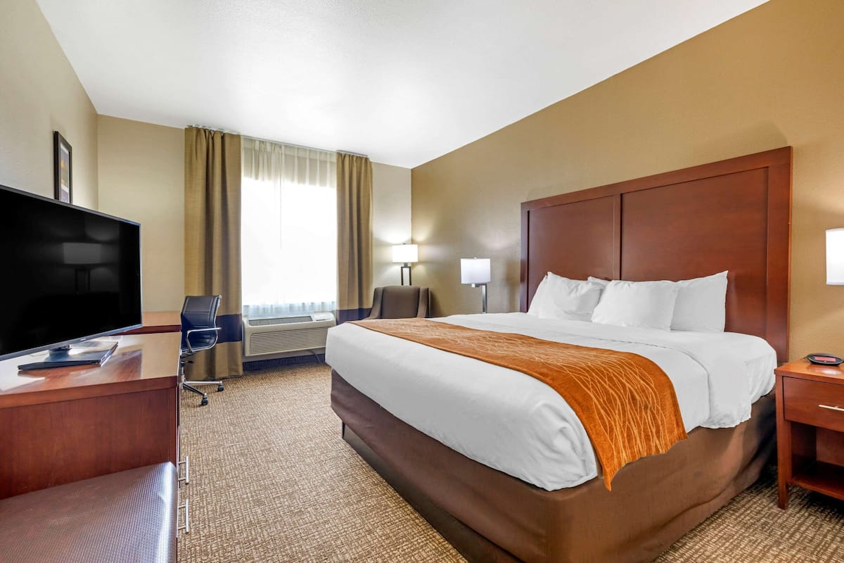 A hotel room with a large bed, a desk with a flat-screen TV, a chair, a lamp, and a nightstand.
