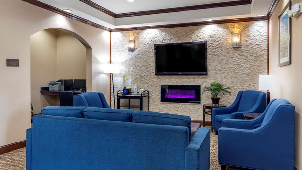 A hotel room with a blue sofa, a blue armchair, a flat-screen TV mounted on a textured wall, a small table with a lamp, and a fireplace. - Comfort Inn Near UNT (Denton)