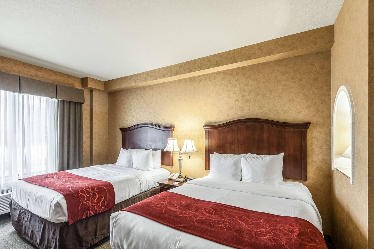 Une chambre d’hôtel avec deux lits, une tête de lit en bois, une table de chevet avec des lampes et une grande fenêtre avec des rideaux.