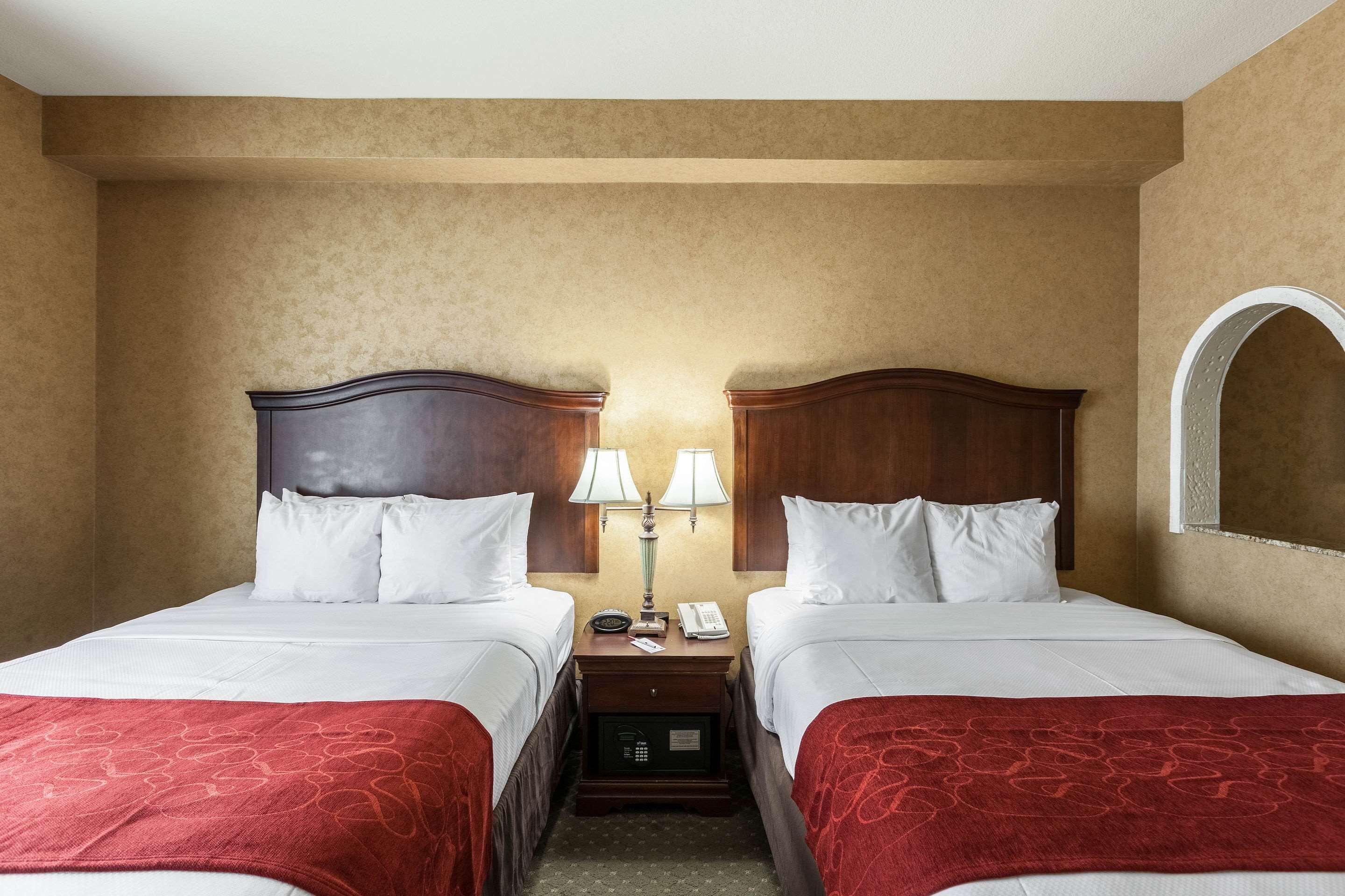 A hotel room with two beds, each with white linens and red throw blankets. There are bedside tables with lamps and a phone. The room has beige walls and a large mirror on one wall.