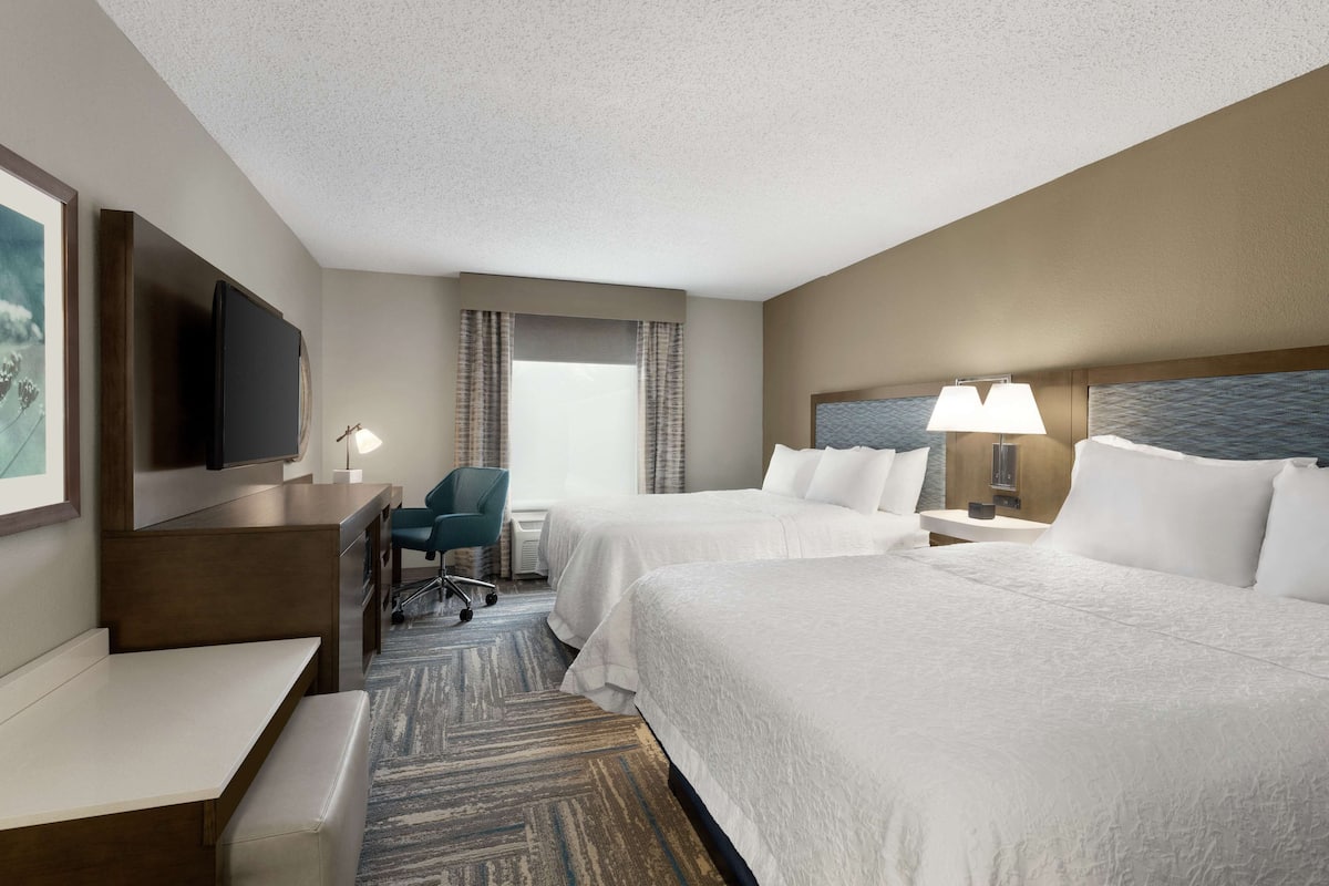 A hotel room with two beds, a desk with a chair, a TV, and a window with curtains.