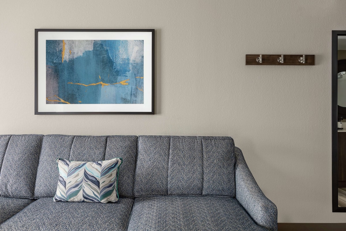 A grey sofa with a patterned cushion, a framed abstract painting, and a wooden shelf with three hooks.