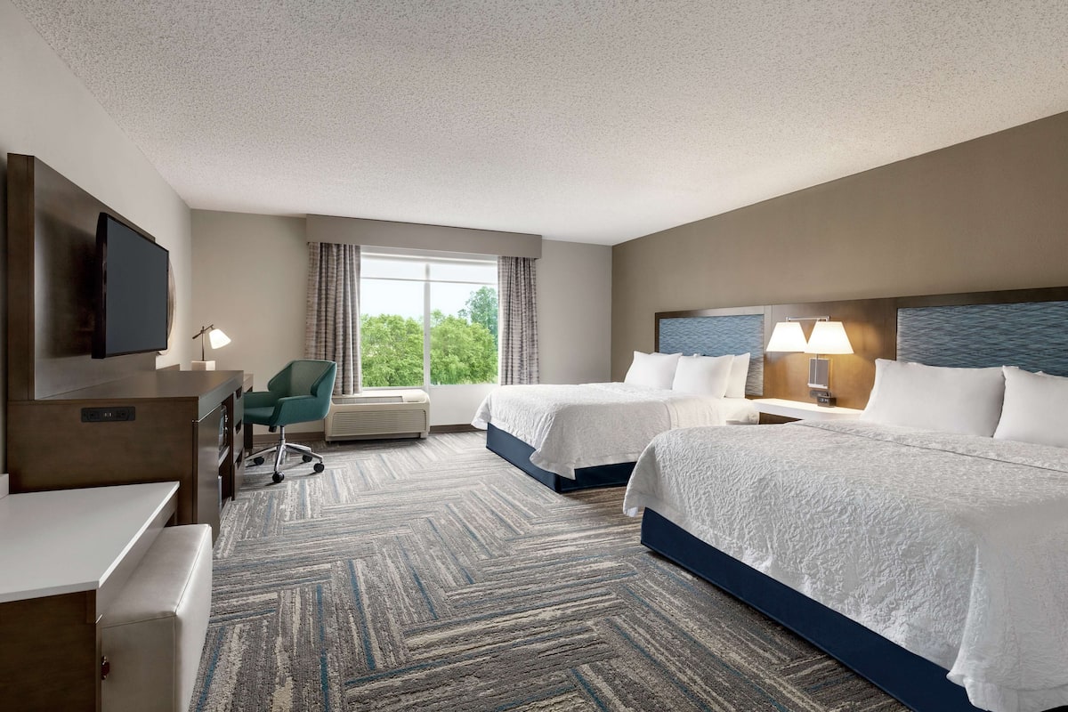 A hotel room with two beds, a desk, a TV, a chair, and a large window with curtains.