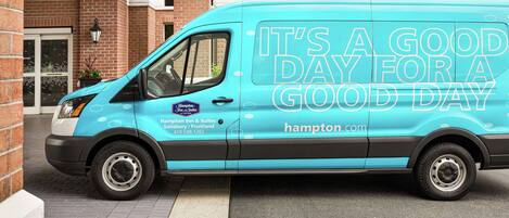 A teal van parked in front of a building with a sign that reads "IT'S A GOOD DAY FOR A GOOD DAY" and the Hampton Inn & Suites logo.
