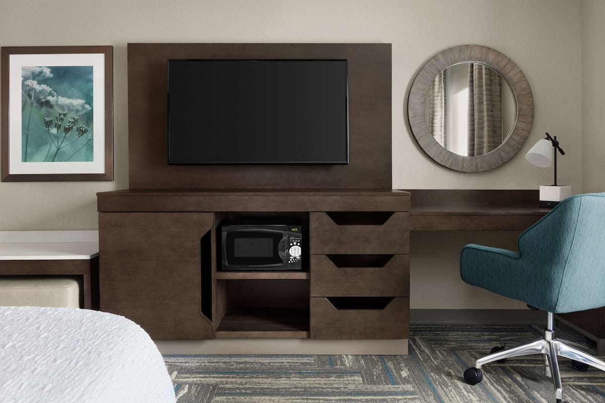A hotel room with a flat-screen TV mounted on a wooden cabinet, a microwave below it, a round mirror above, and a teal office chair.