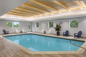 An indoor swimming pool with wooden deck flooring, multiple windows, and Adirondack chairs.
