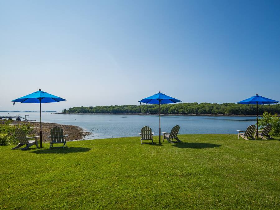 A grassy area with three Adirondack chairs and blue umbrellas, overlooking a body of water and a distant shoreline.