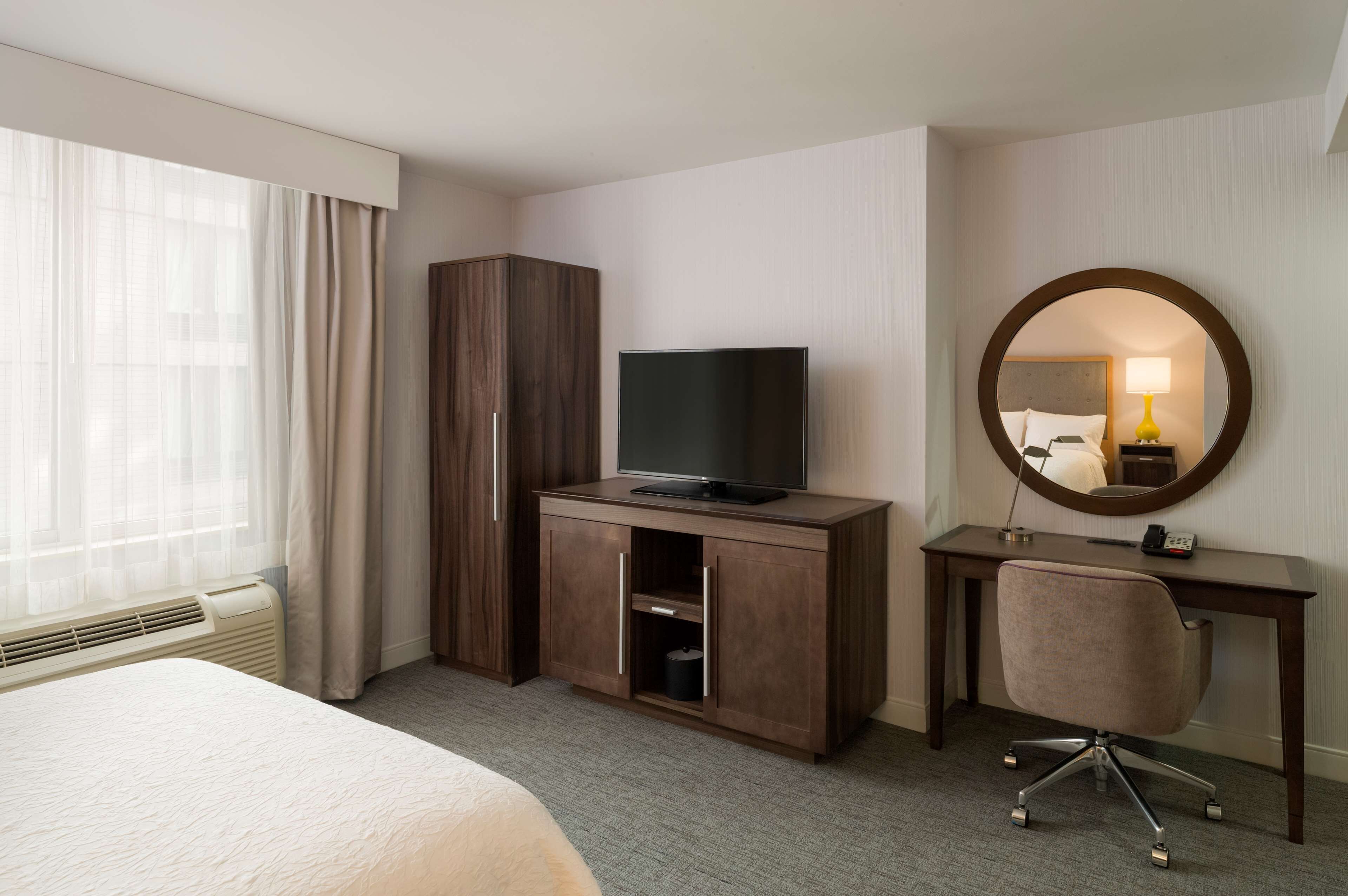 A hotel room with a bed, a TV on a wooden cabinet, a desk with a chair, and a large mirror.