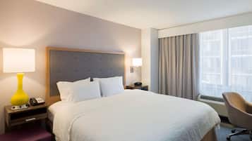 Quarto de hotel com uma cama grande, uma mesinha de cabeceira com luminária, uma cadeira e uma janela com cortinas.