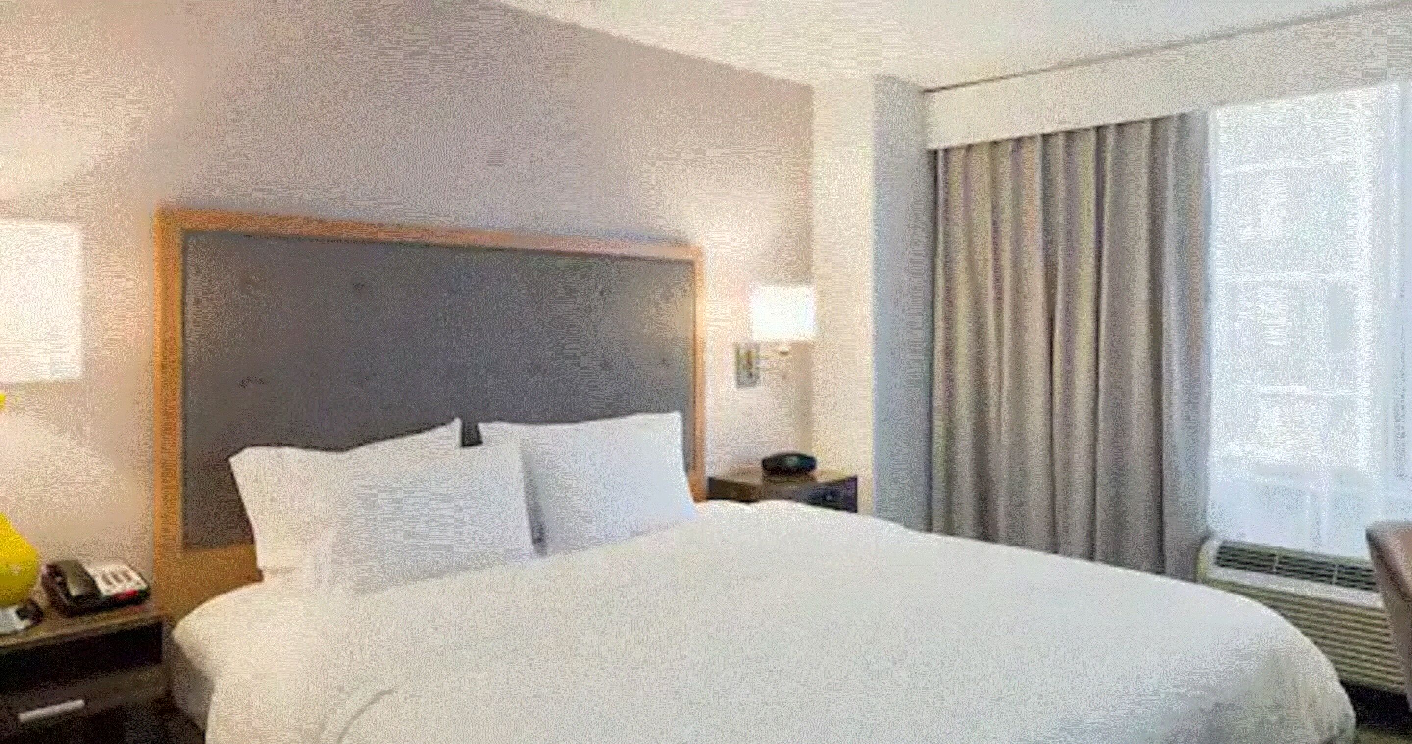 A hotel room with a large bed, a nightstand with a lamp, a bedside table, and a window with curtains.