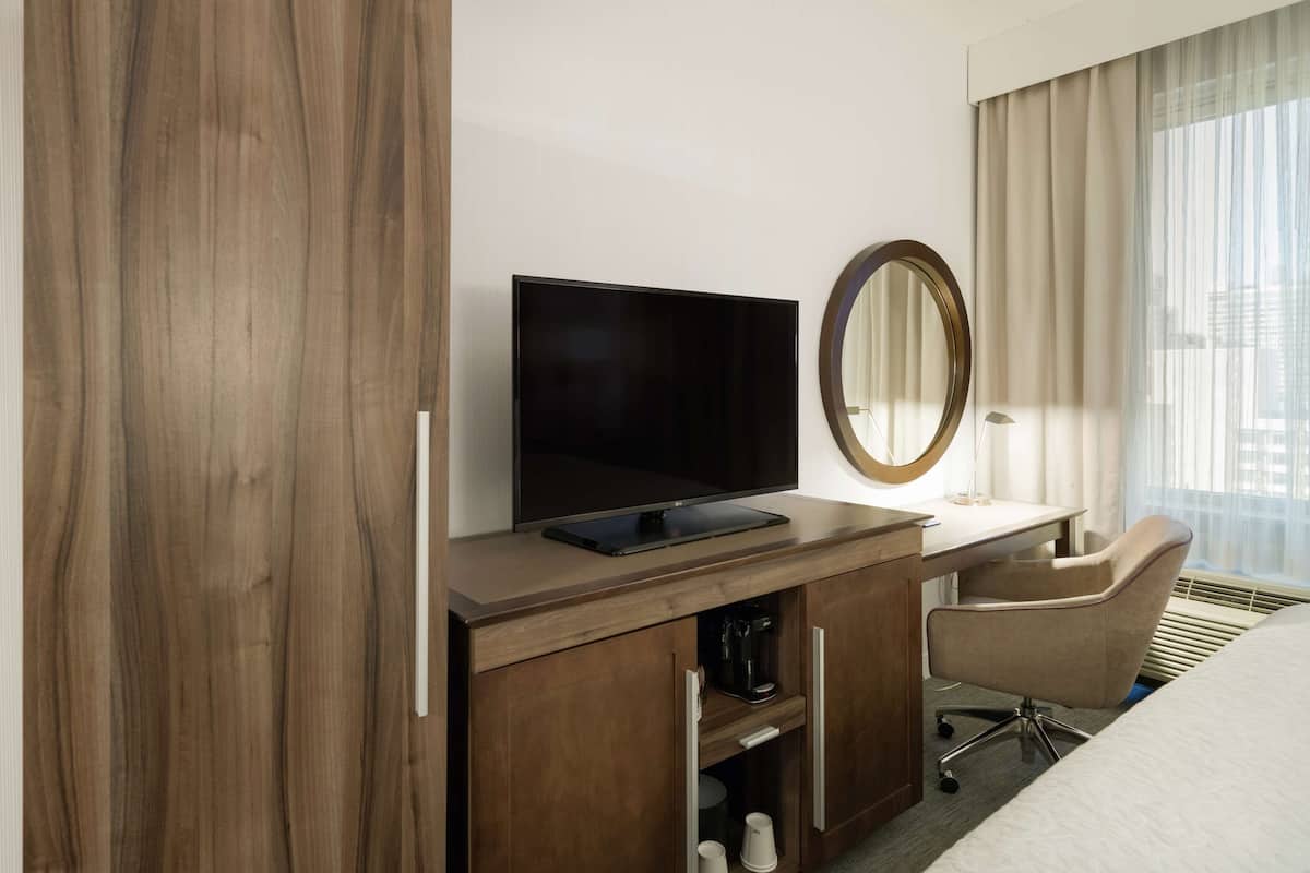 A hotel room with a wooden desk, a flat-screen TV, a coffee maker, a chair, and a large window with curtains.