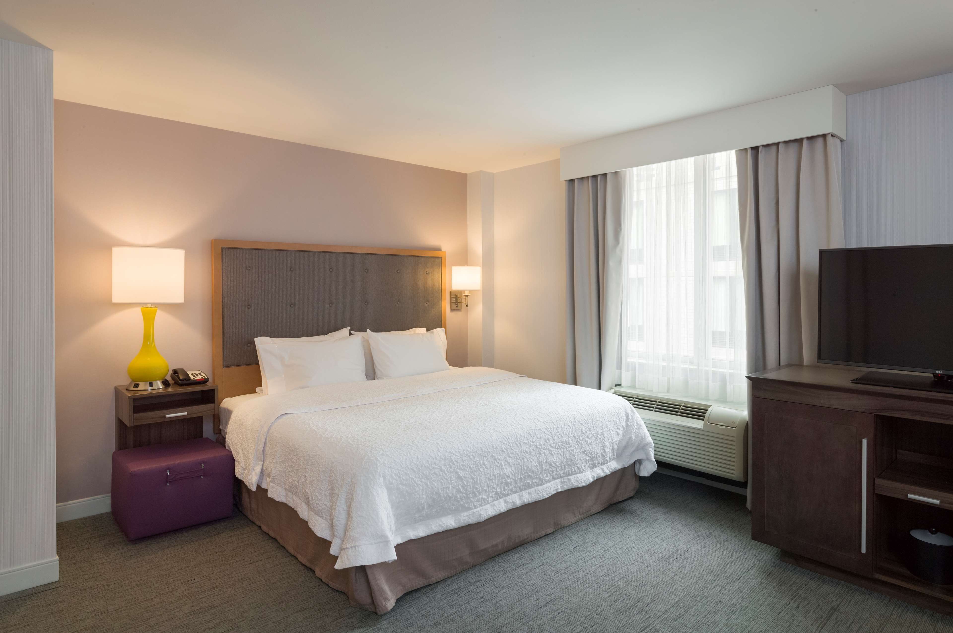 A hotel room with a bed, a nightstand with a lamp, a suitcase, a television, and a window with curtains.