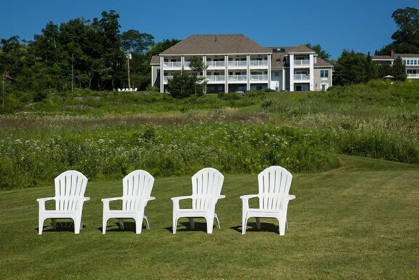 A building with multiple floors and balconies, surrounded by greenery and a grassy area with four white chairs.