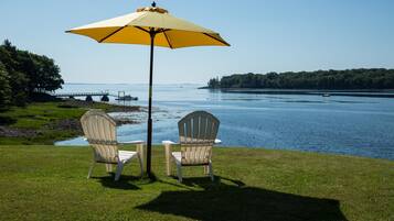 Two Adirondack chairs under a yellow umbrella on a grassy area with a lake view.