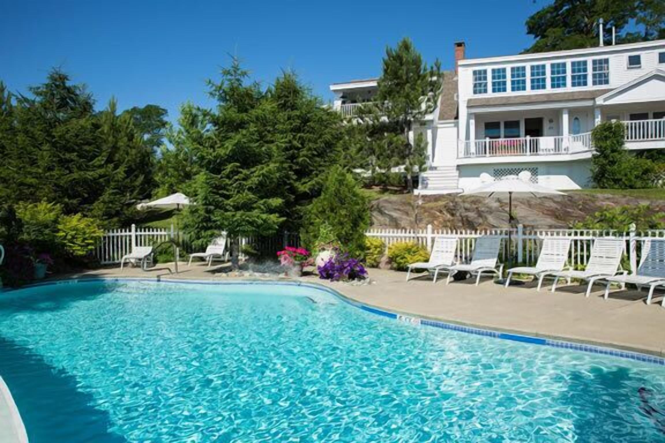 Une piscine avec des chaises longues et une clôture blanche, entourée d’arbres et d’un bâtiment doté d’un balcon.