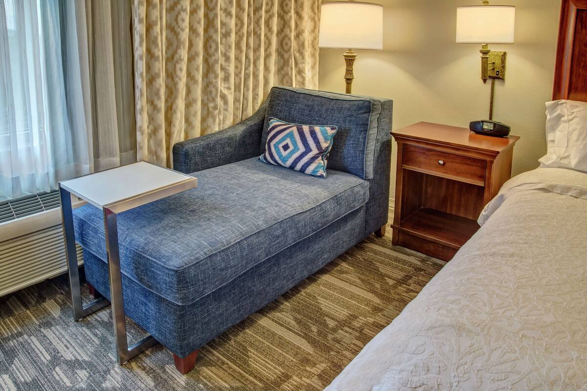 Une chambre d’hôtel avec un canapé bleu, une petite table, une table de chevet en bois, une lampe et un lit.