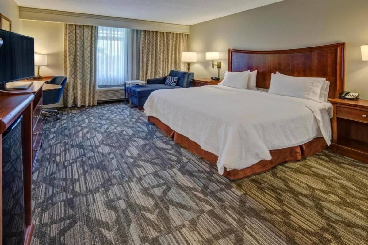 A hotel room with a large bed, a desk, a chair, a TV, and a window with curtains.