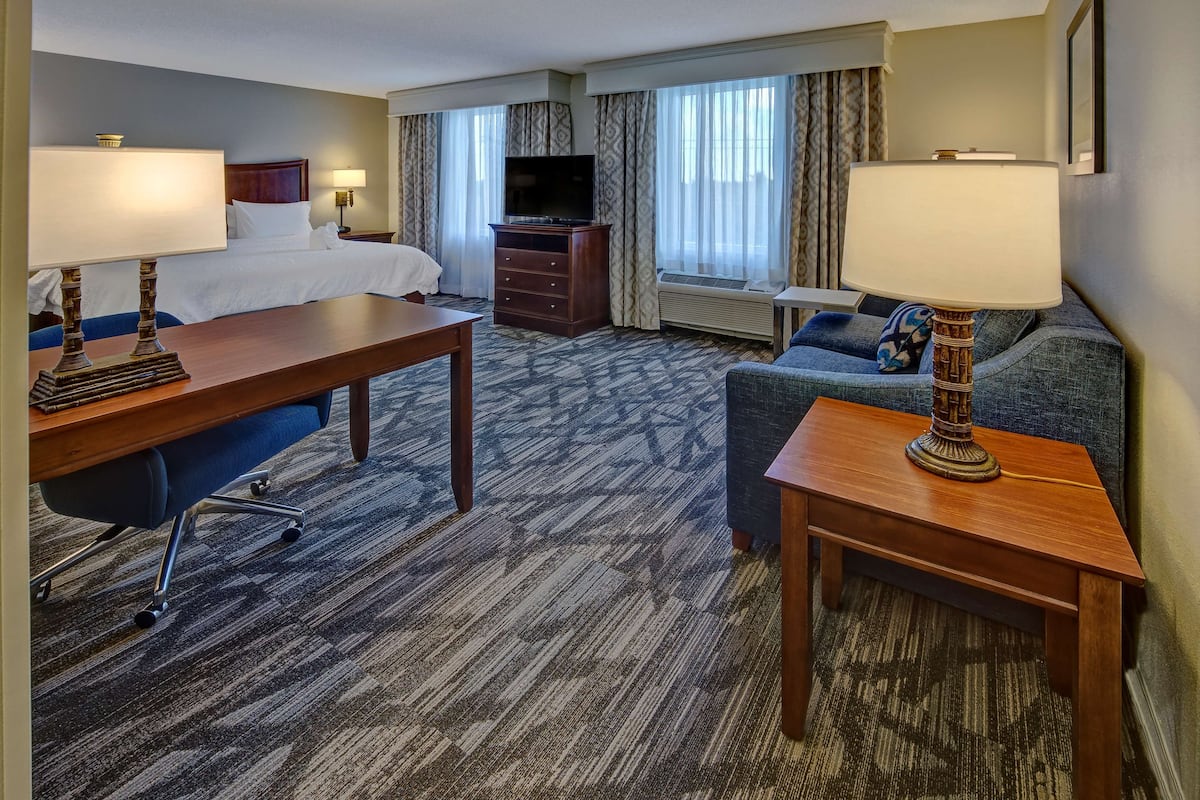 A hotel room with a bed, a desk with a chair, a sofa, a TV on a dresser, and a window with curtains.