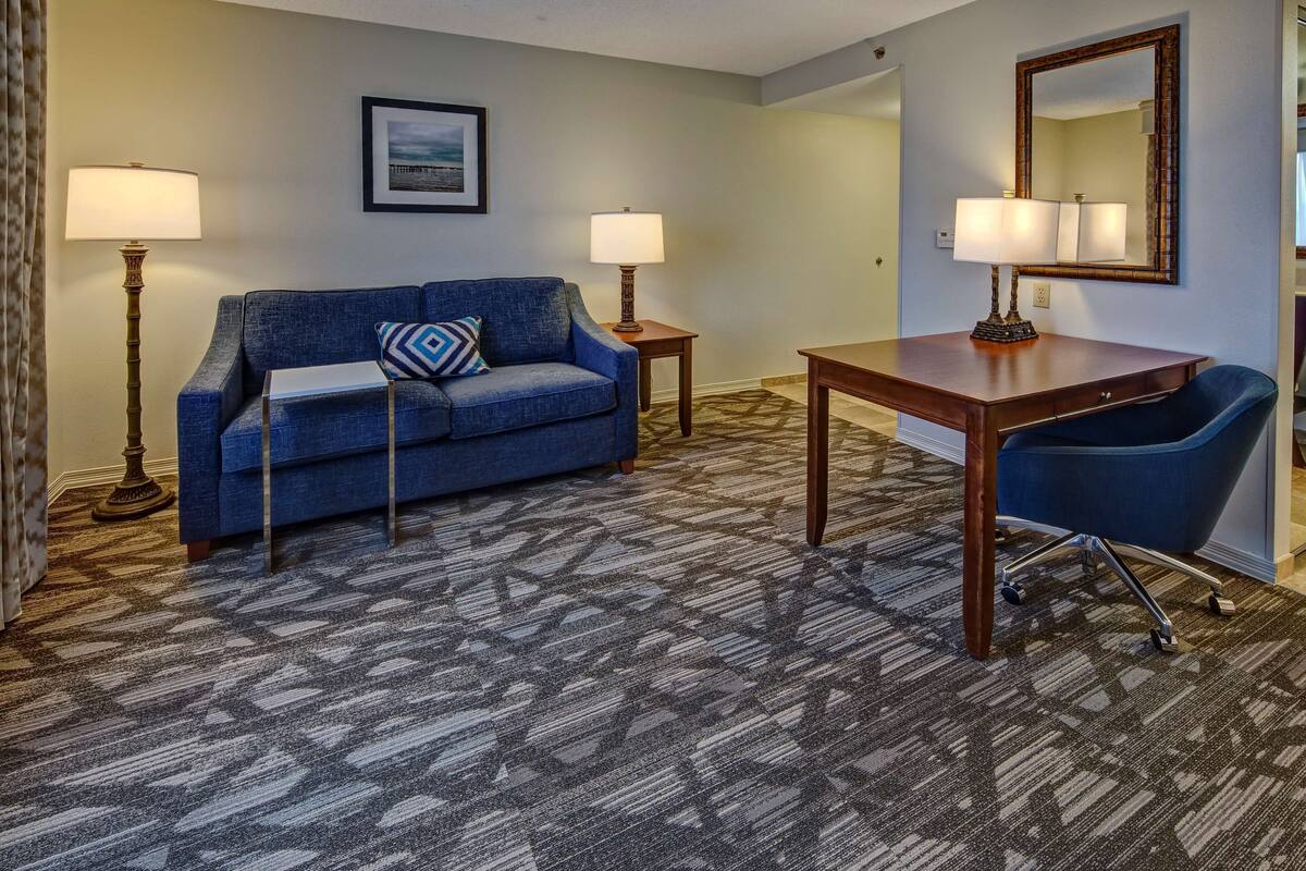 A hotel room with a blue sofa, a wooden desk, two lamps, and a patterned carpet.