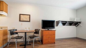 Una habitación pequeña con una mesa de madera, dos sillas, un televisor, un mueble bajo y un estante en la pared con una cesta.