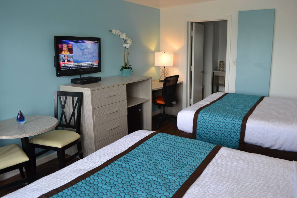 Ein Hotelzimmer mit zwei Betten, einem Schreibtisch, einem Fernseher, einem Sessel, einer Lampe und einem kleinen Tisch.