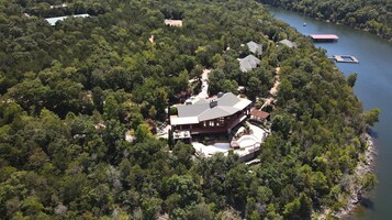 A large, single-story house surrounded by dense forest, with a dock extending into a lake.