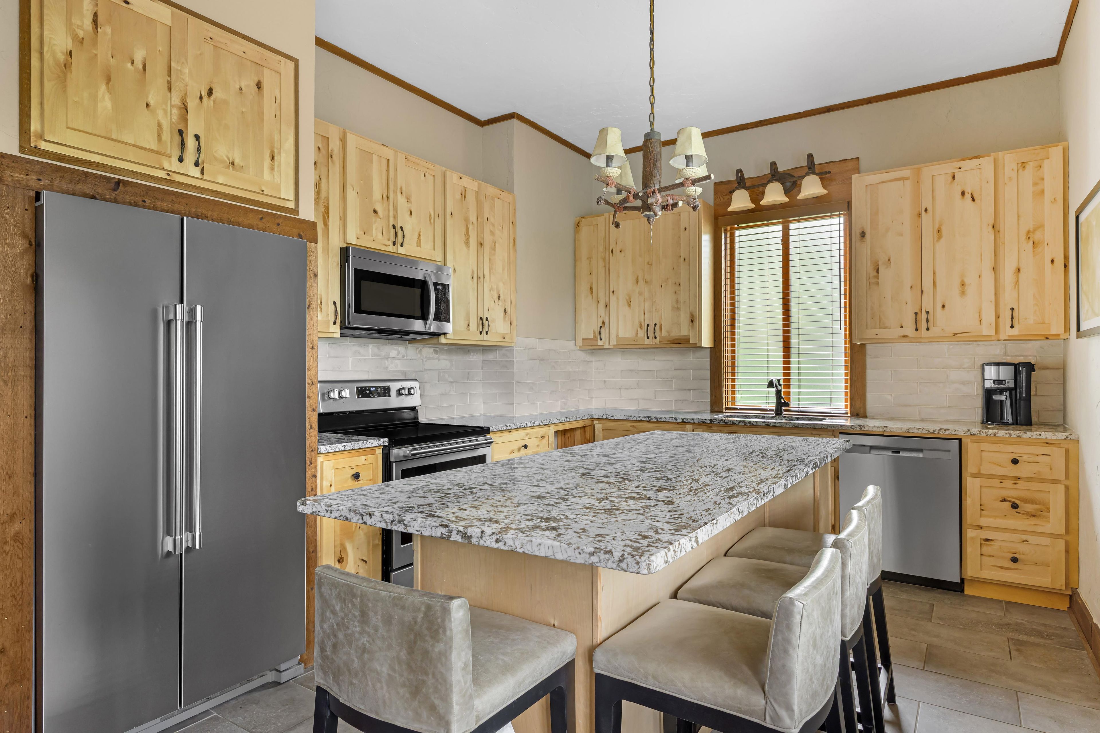 A kitchen with wooden cabinets, a stainless steel refrigerator, a microwave, a stove, a sink, and a marble countertop.