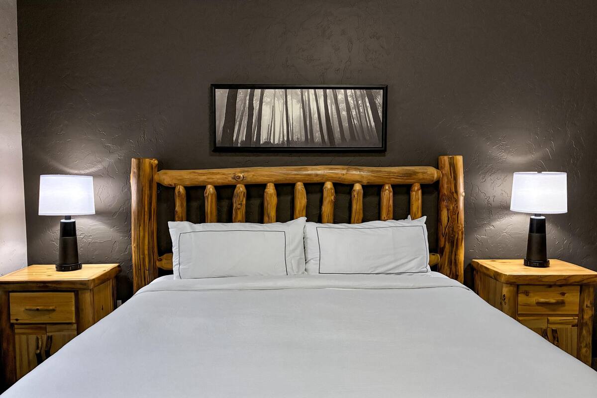 Una cama con cabecera de madera, dos mesitas de noche con lámparas, y un cuadro enmarcado en la pared.