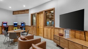 A room with a large flat-screen TV, wooden paneling, a table with chairs, and arcade games.