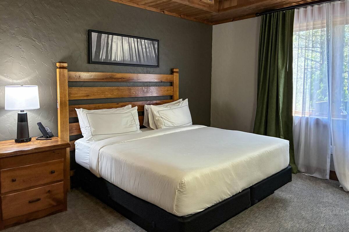 Un dormitorio con cabecera de madera, cama con ropa de cama blanca, mesita de noche con lámpara y teléfono, y ventana con cortinas verdes.