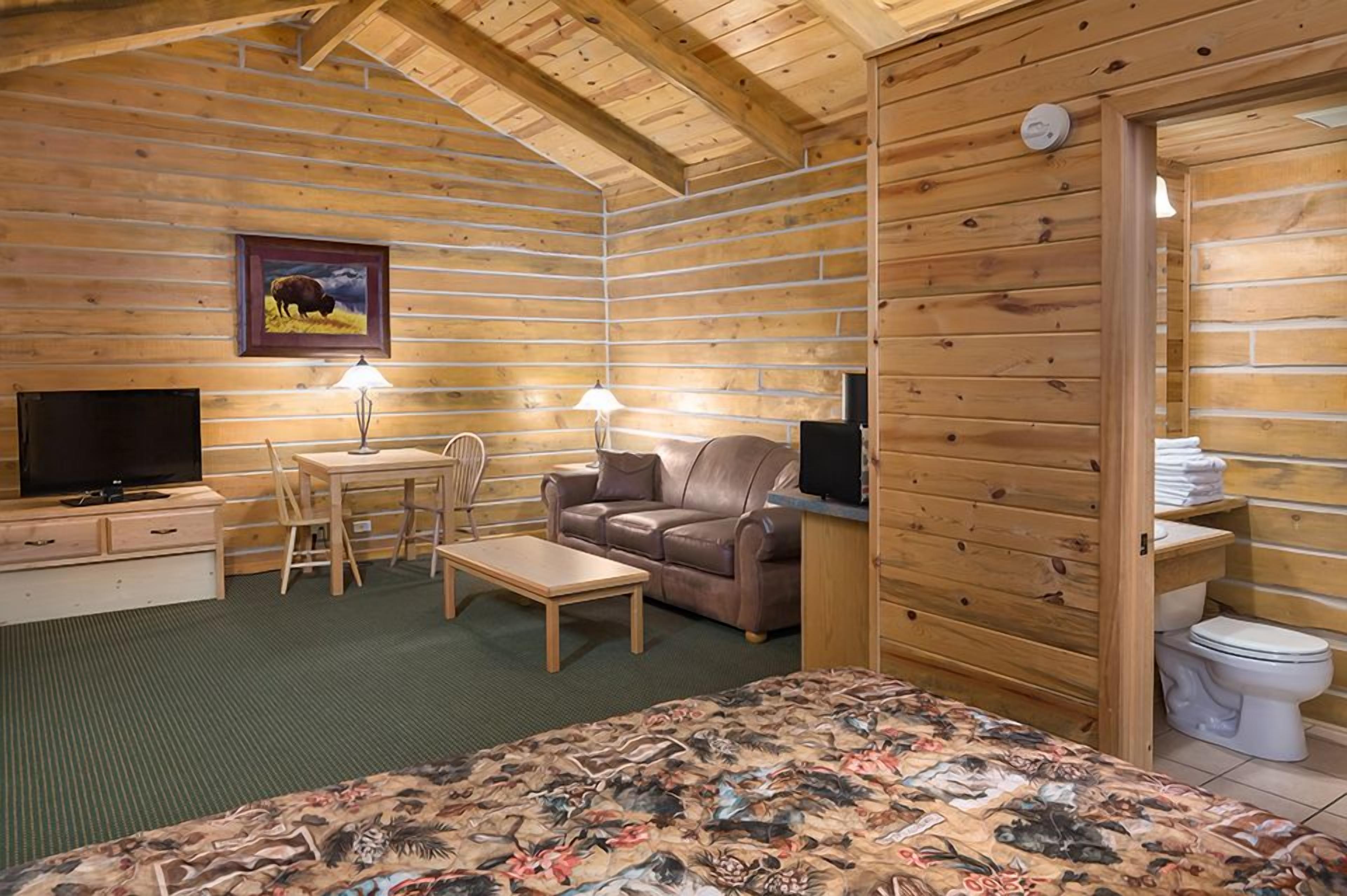 A log cabin room with a bed, a sofa, a television, and a desk with a lamp.