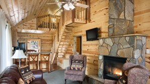 A log cabin interior with a stone fireplace, a wooden staircase, a leather sofa, and a television mounted on the wall. - Rock Crest Lodge And Cabins (Custer)