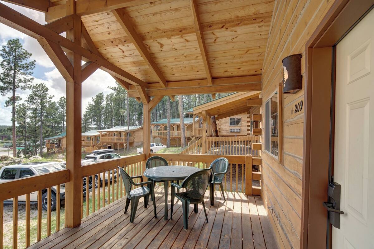 Un terrazzo in legno con tavolo e sedie, affacciato su una zona boschiva con diverse cabine.