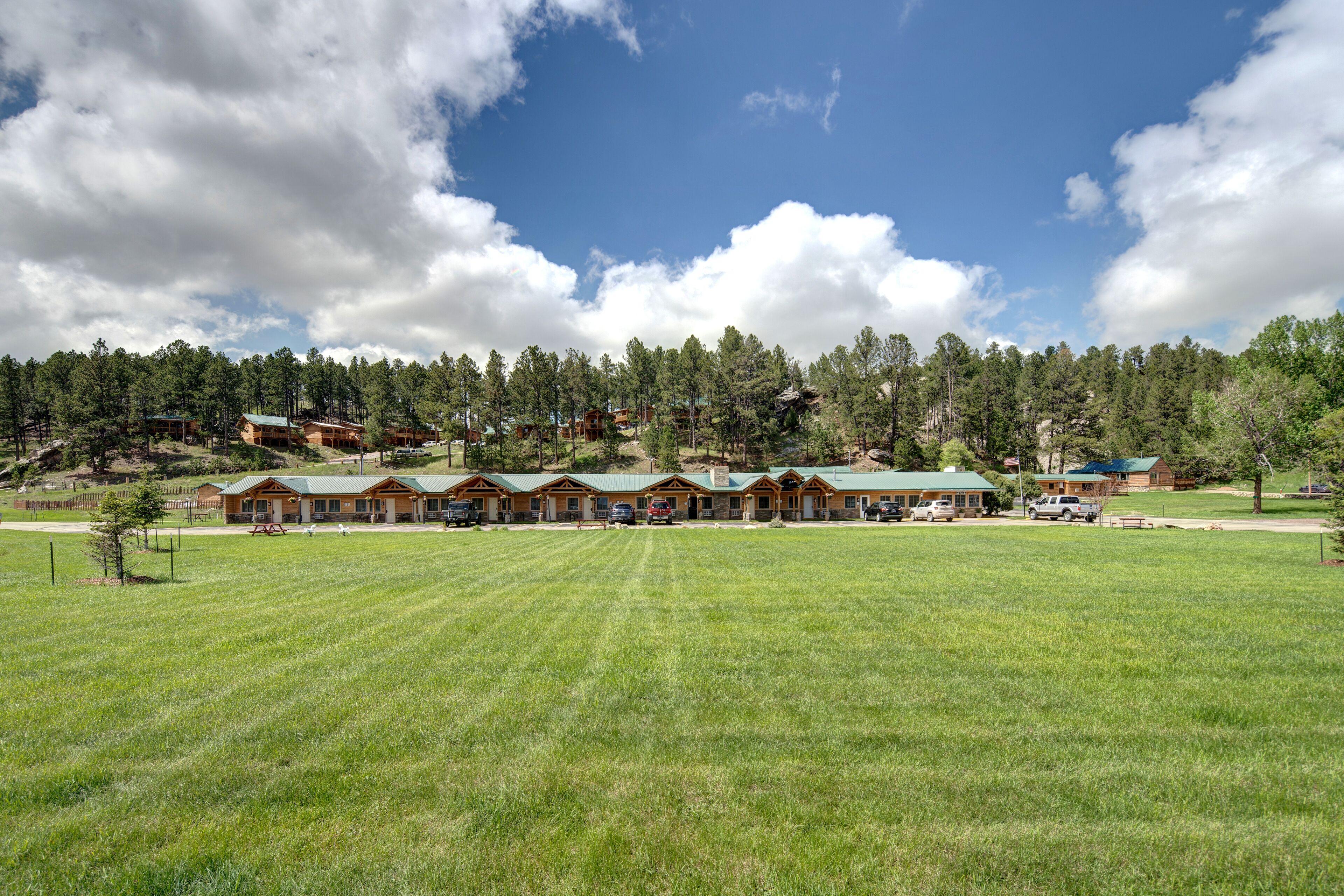 A lodge with multiple cabins, a large grassy field, and a forested hillside in the background.