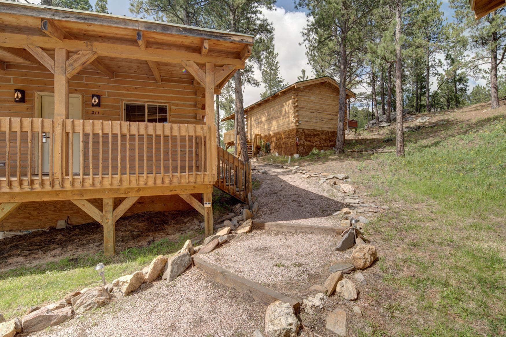 A wooden cabin with a porch, a gravel path, and a forested hillside.