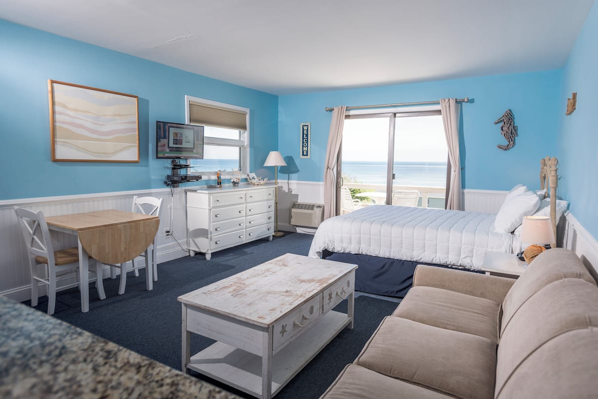 A room with a bed, a desk, a chair, a coffee table, and a view of the sea.