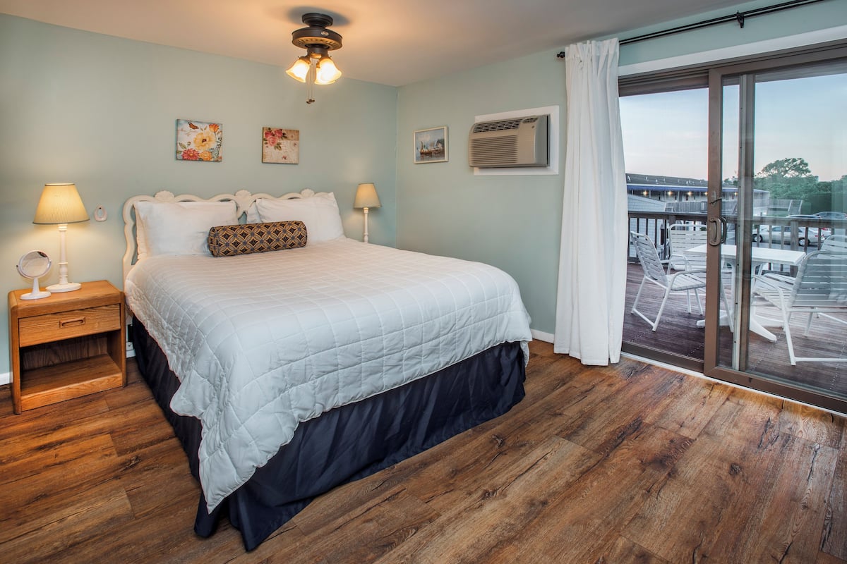 A bedroom with a large bed, wooden flooring, a ceiling fan, and a balcony with outdoor furniture.