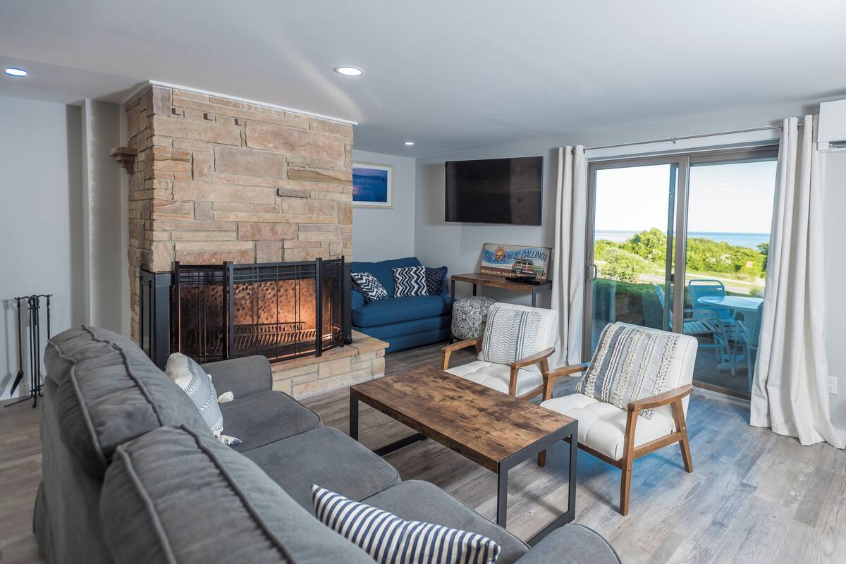A living room with a stone fireplace, a sofa, a coffee table, and a view of the outdoors.