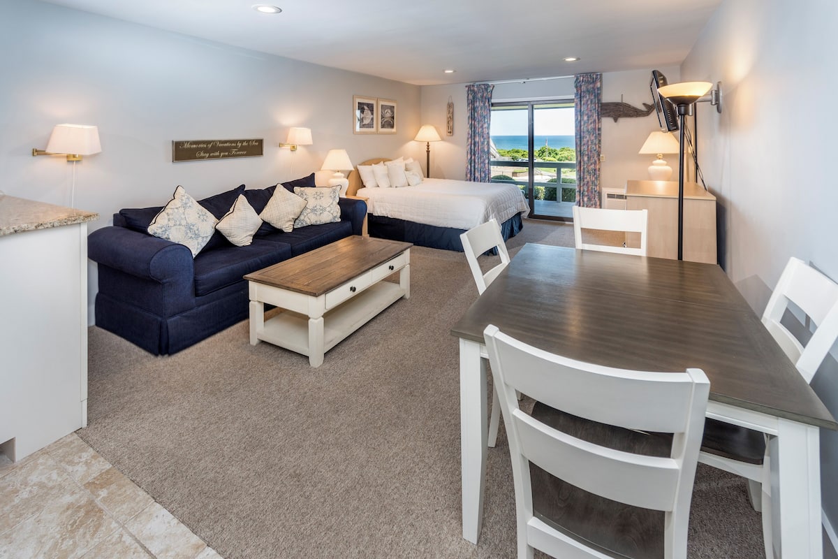 A hotel room with a bed, a sofa, a dining table, and a view of the sea.