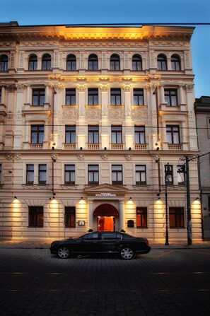 Ein historisches Gebäude mit einer klassischen Fassade und einem markanten Eingang. Vor dem Gebäude ist ein schwarzes Auto geparkt.