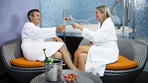 Two people in white robes seated on wicker chairs with orange cushions, toasting with champagne glasses.