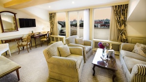 A spacious living room with striped sofas, a dining area with a table and chairs, a large window with a city view, and a flat-screen TV mounted on the wall.
