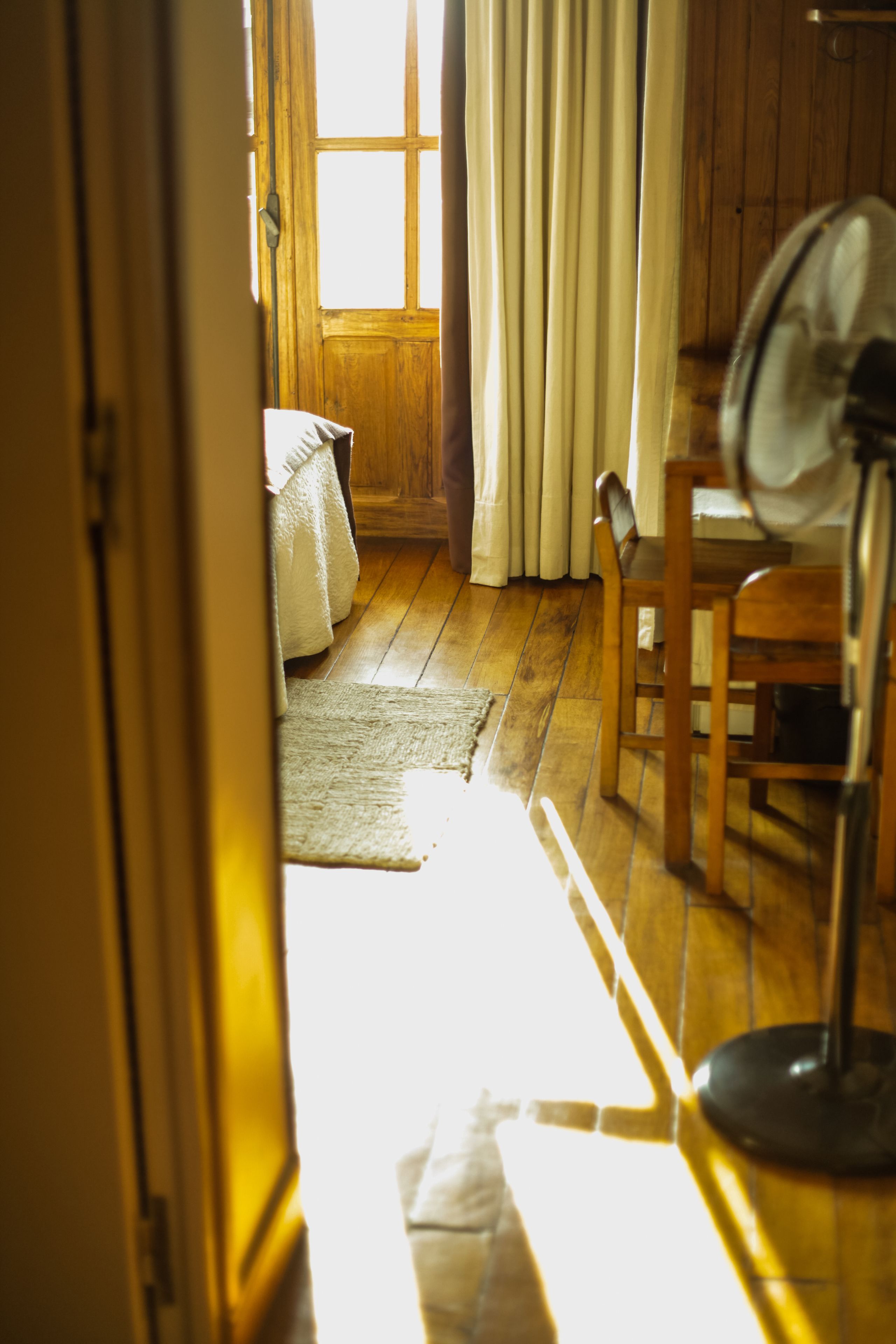 A room with wooden flooring, a bed with white linens, a wooden chair, a table, and a standing fan.