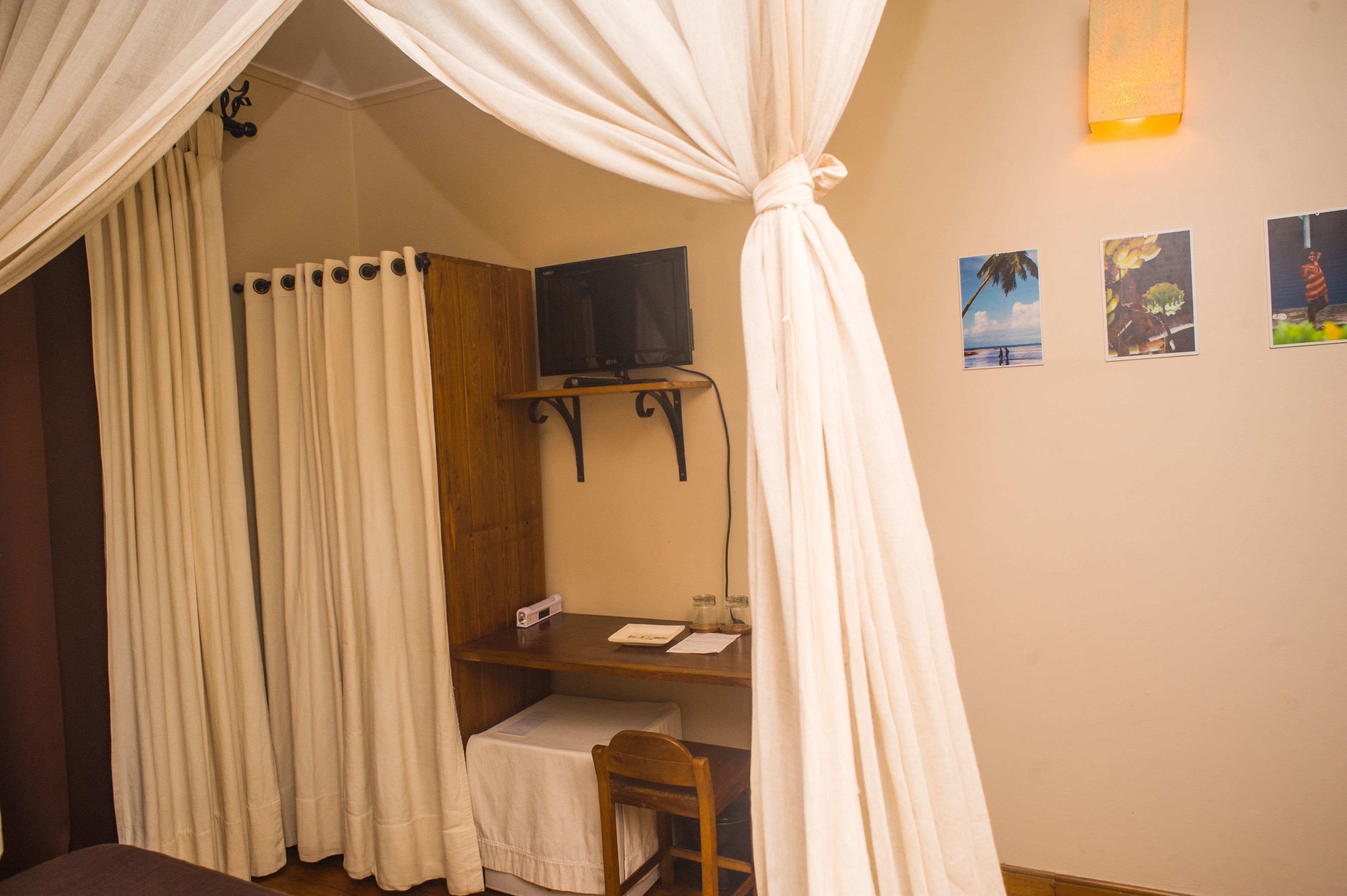 A room with a bed covered by white curtains, a desk with a television, and wall-mounted pictures.