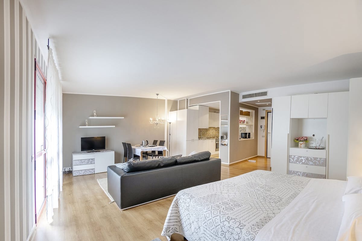 A modern bedroom with a large bed, a sofa, a TV, and a kitchen area in the background.